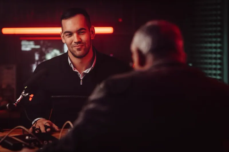 Studio: Mann im schwarzen Pullover am Tisch mit Mikrofon, Standbild aus dem Video