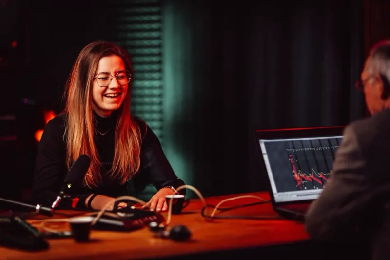 Studio: lachende Frau mit Brille und langen Haaren am Tisch mit Mikrofon, Standbild aus dem Video