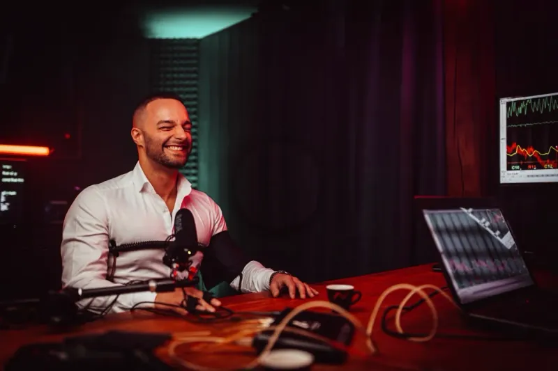 Studio mit rot-grünem Licht: lachender Mann im weißen Hemd mit Messgurten am Tisch mit Laptop und Mikrofon (Pierre Martins Rodrigues)