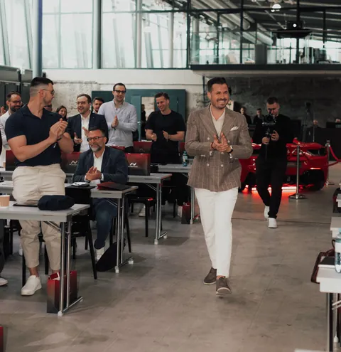Nino Fischlein im karierten Sakko geht applaudierend durch einen Seminarraum, Teilnehmer an Tischen klatschen, im Hintergrund ein rotes Sportauto