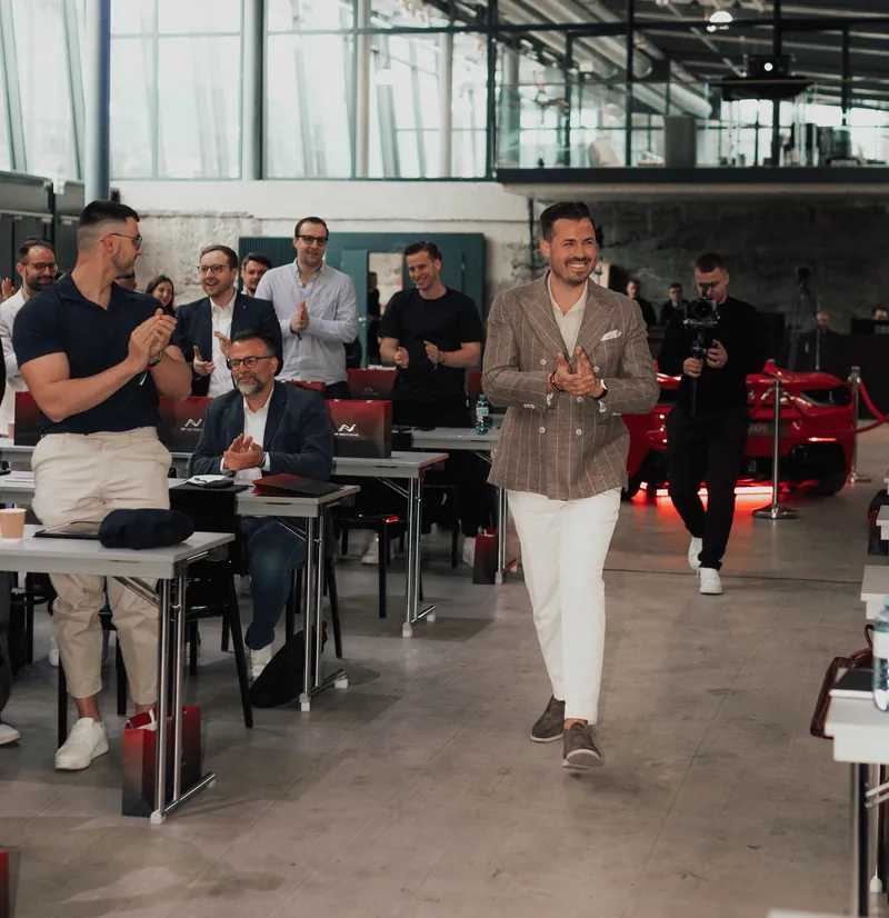 Nino Fischlein im karierten Sakko geht applaudierend durch einen Seminarraum, Teilnehmer an Tischen klatschen, im Hintergrund ein rotes Sportauto