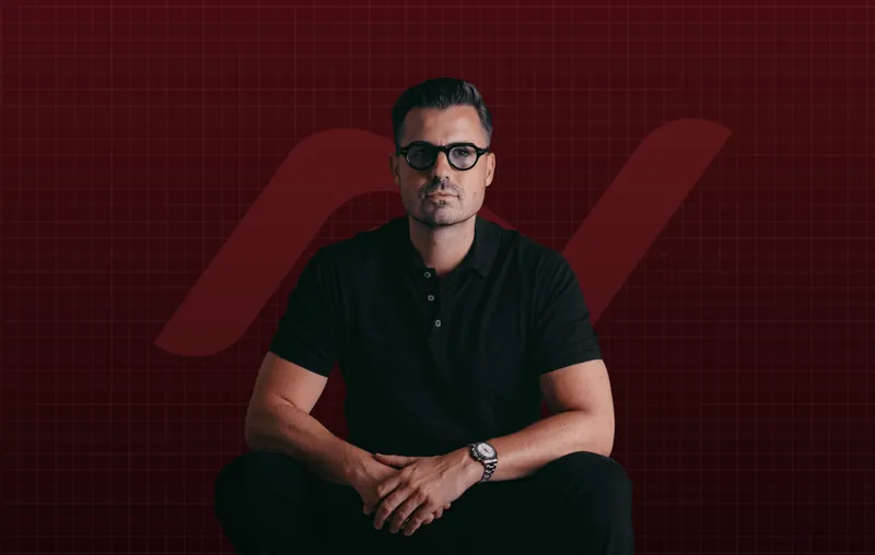 Nino Fischlein sitzend, schwarzes Poloshirt, Brille und Armbanduhr, vor dunkelrotem Hintergrund mit dem NF-Signet