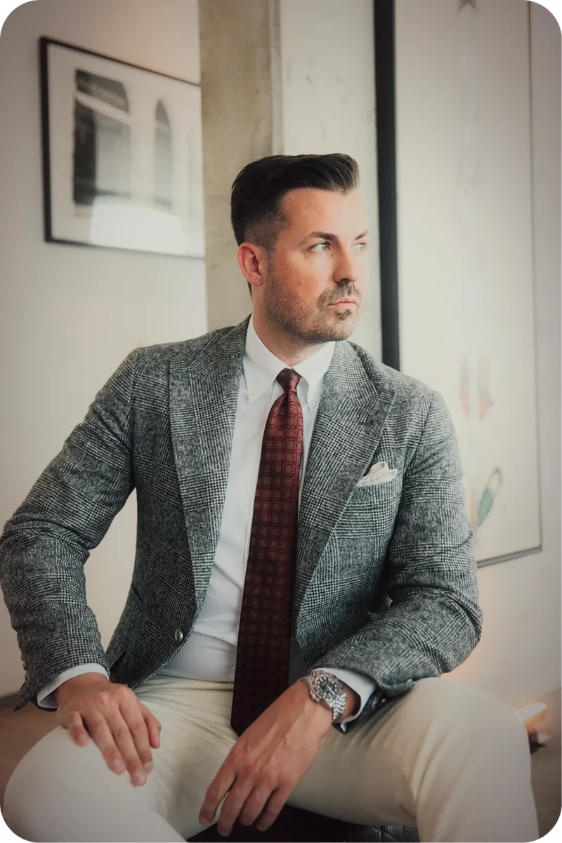 Nino Fischlein sitzend im grauen Tweed-Anzug mit weinroter Krawatte in einem Büro mit Bildern an der Wand